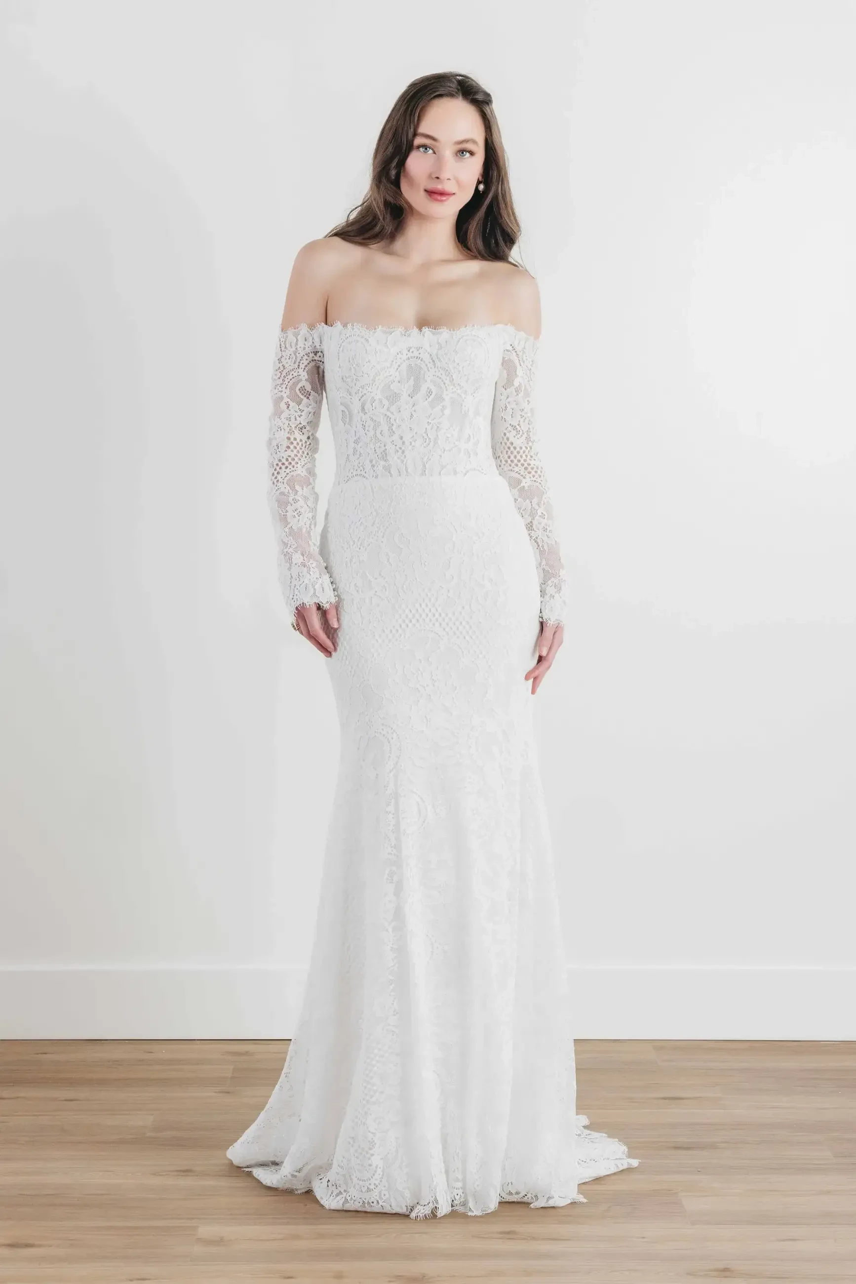 Model is wearing a fitted, off-the-shoulder lace gown with long sleeves and a low illusion back, perfect for a romantic fall or winter wedding.