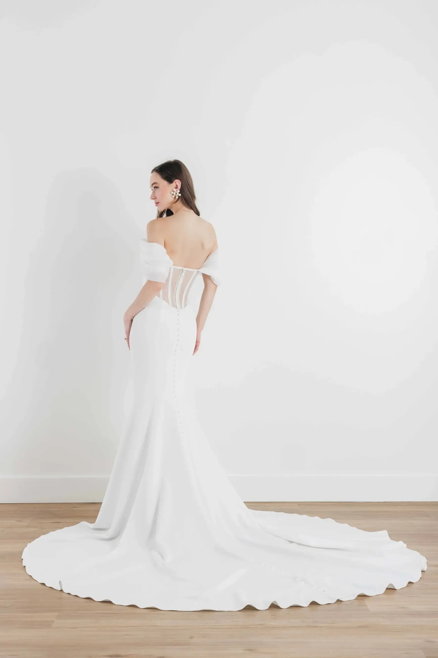 Model is wearing a fit and flare off the shoulder wedding dress with ruching on the bodice and long train