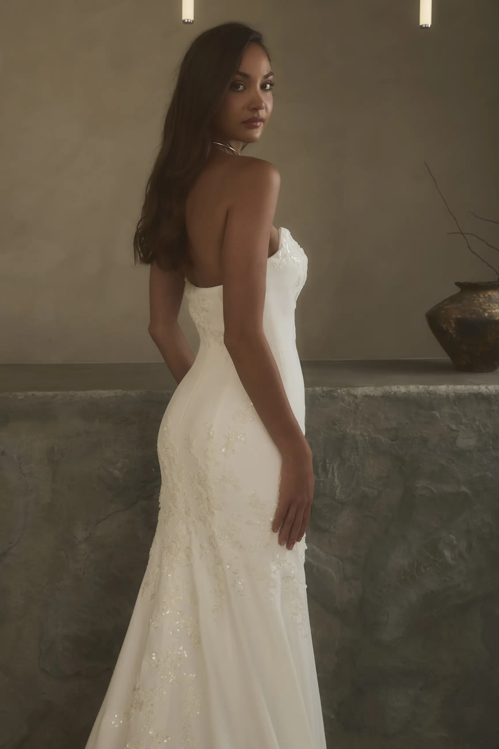 Elegant woman in a strapless, white gown with subtle floral details stands by a textured, stone-like wall. The setting is minimal and serene.