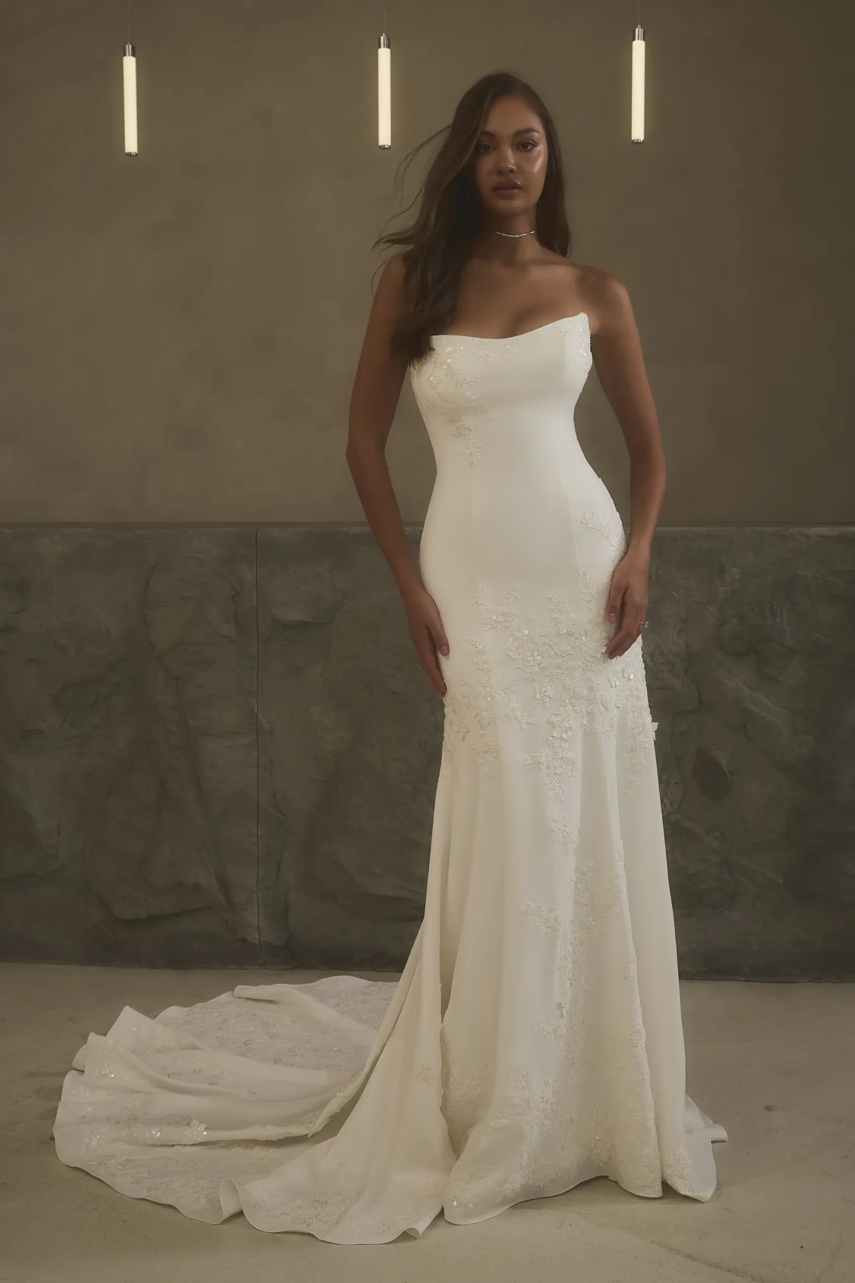 A woman in an elegant, strapless white wedding gown with subtle lace detailing stands in a sophisticated setting, illuminated by three vertical lights.