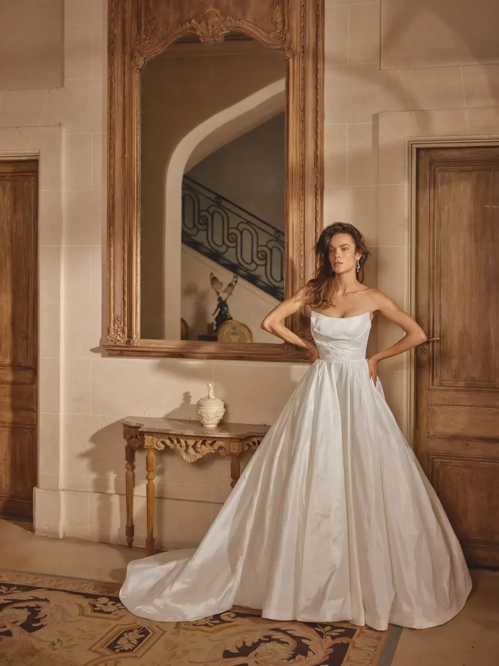 A model poses in a flowing white gown in an elegant interior, featuring ornate mirrors and wooden doors.