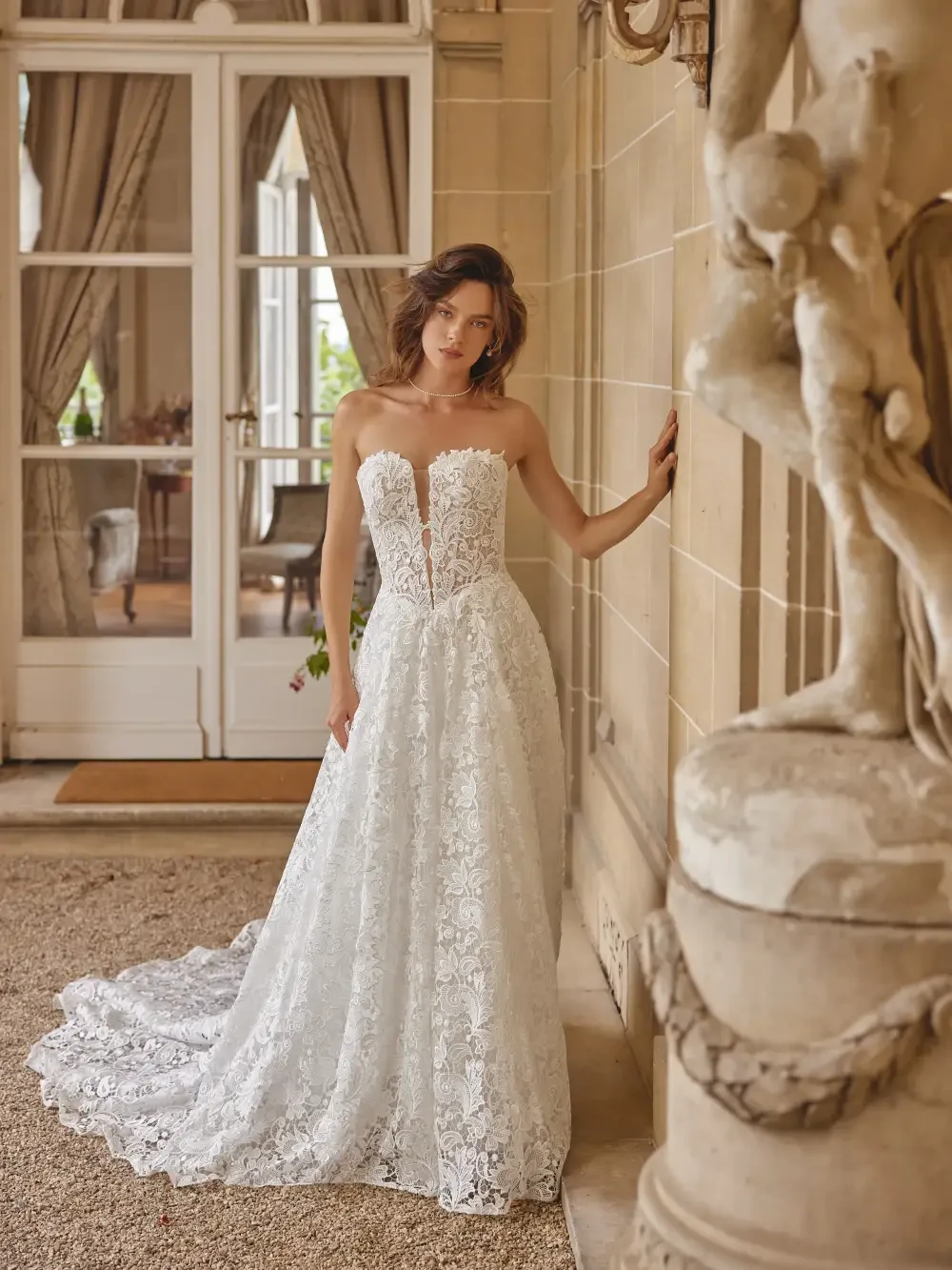 A woman in a floral lace wedding dress stands by a stone wall, leaning against it, with a soft expression. The background features elegant decor and light streaming in through large windows.