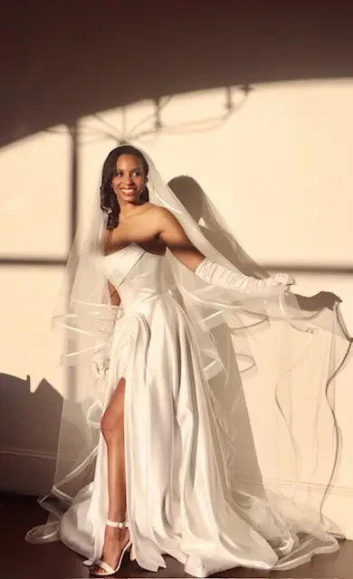 Bride in a flowing white dress stands in warm sunlight, casting elegant shadows. She smiles, exuding joy and grace, with a veil and gloves completing her look.