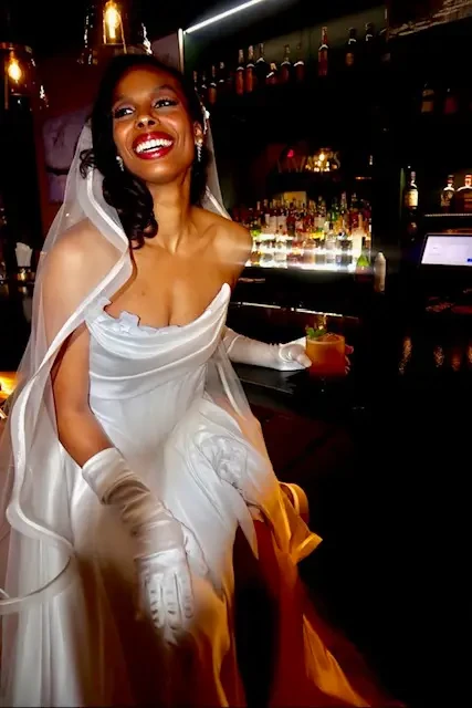 Smiling bride in elegant gown and gloves, holding a drink at a dimly lit bar. Bottles and soft lighting in background create a joyful, celebratory atmosphere.