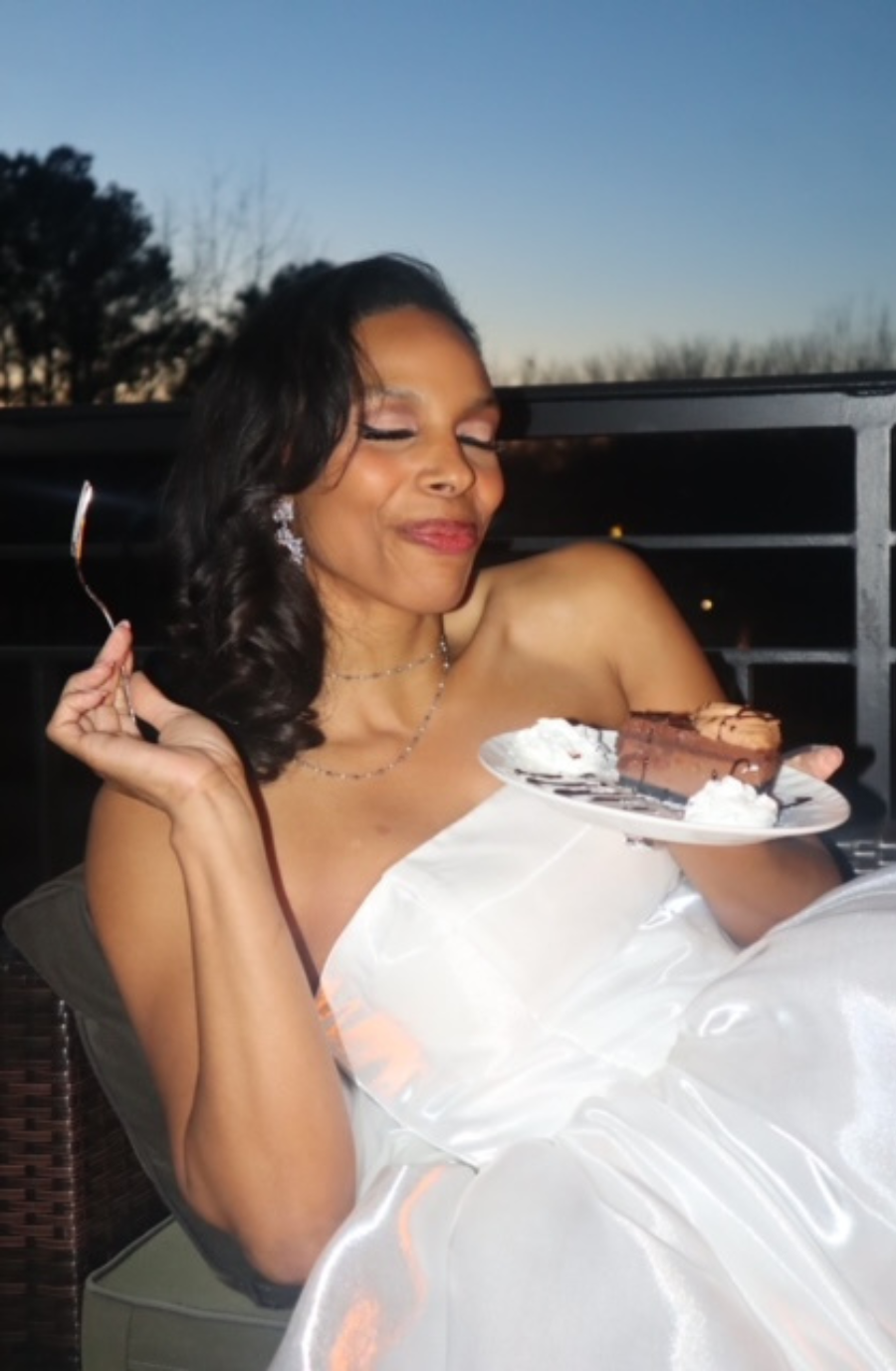 A woman in a white strapless dress holds a fork and a dessert plate with chocolate cake and whipped cream. She smiles contentedly, with a twilight sky behind her.