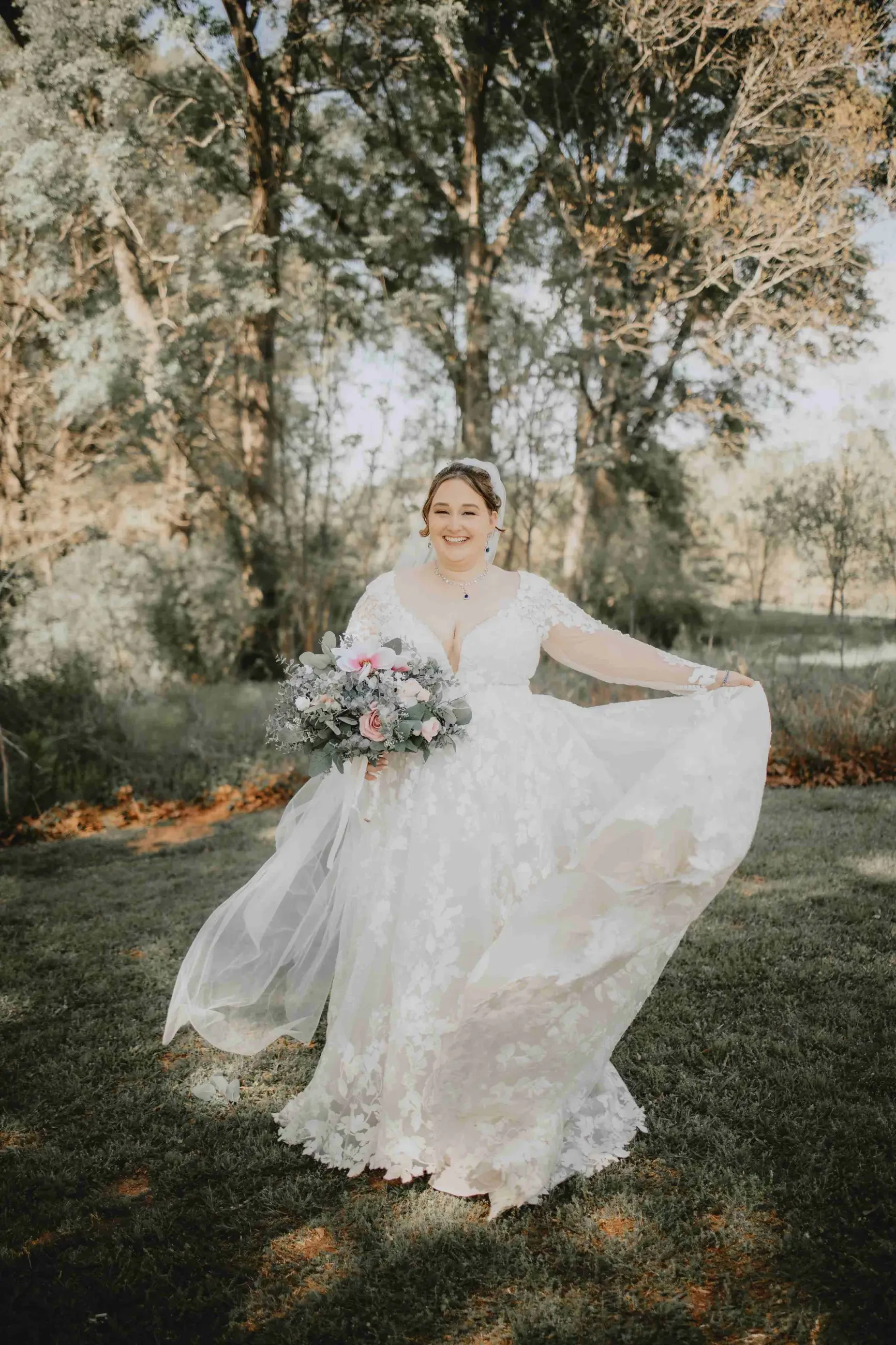 A joyful bride in a flowing lace wedding gown holds a bouquet with pastel flowers. She stands on lush grass, with tall trees in the serene background.