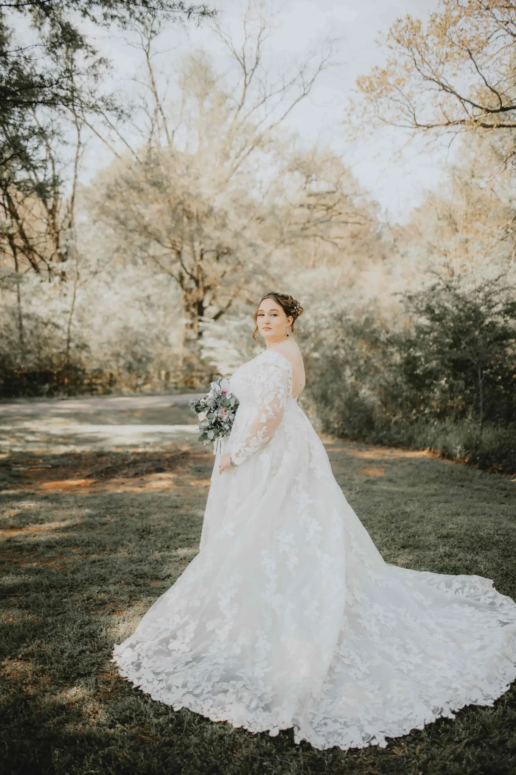 A bride in a flowing lace gown holds a bouquet, standing on lush green grass with a backdrop of sunlit trees. The scene is serene and romantic.