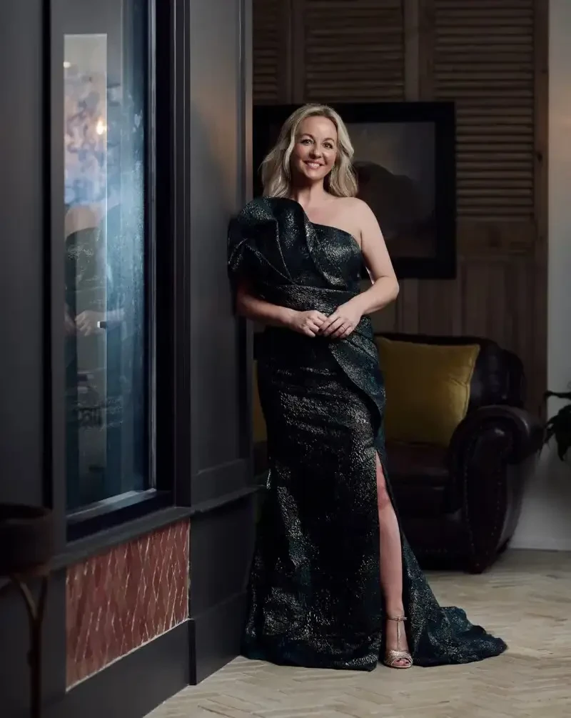 A woman wearing a long black dress with one-shoulder sleeves, standing against a black background.