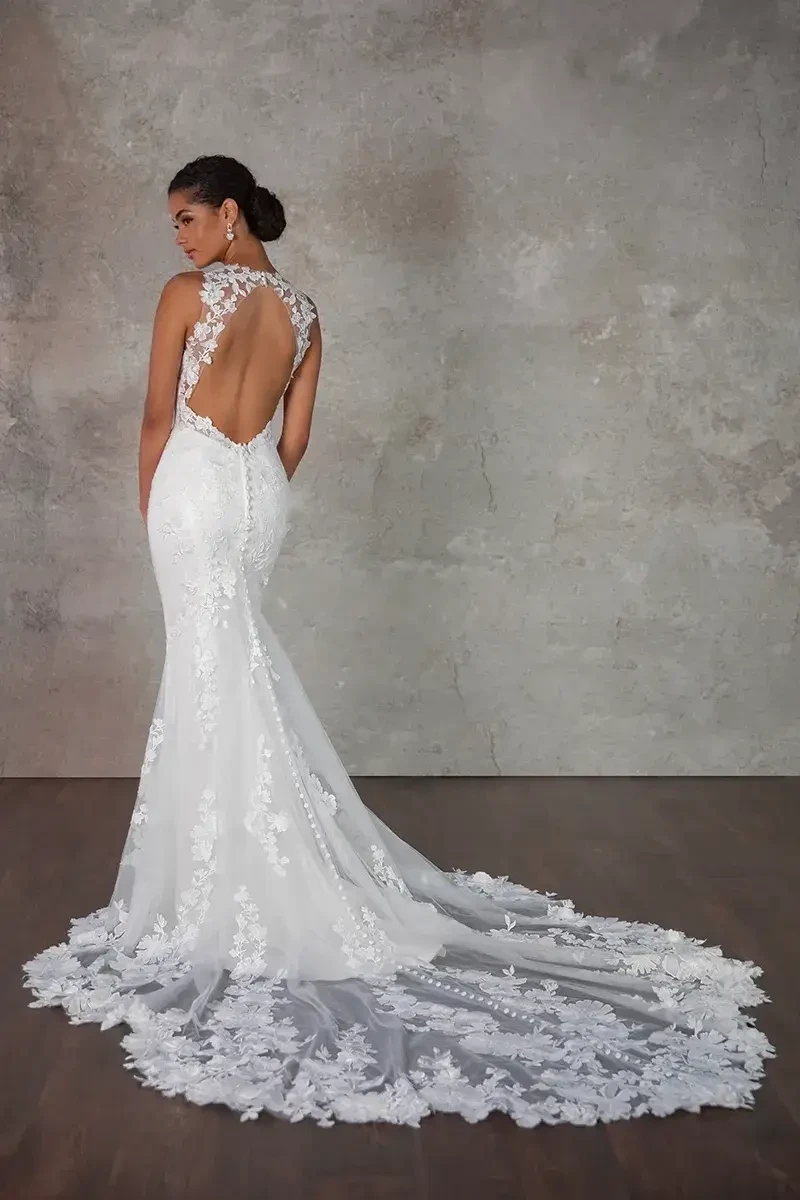 A woman in an elegant, backless white lace wedding gown stands on a wooden floor. The dress has a long, flowing train with floral patterns.