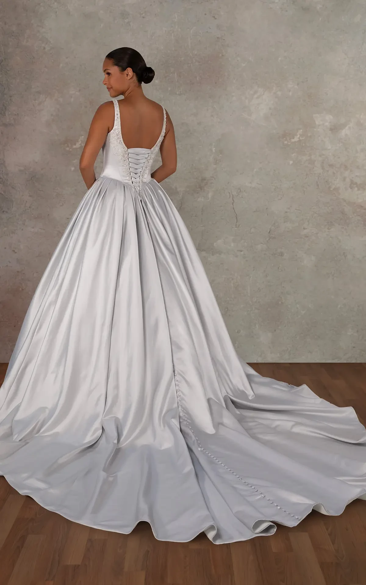 A woman in a satin wedding gown stands on a wooden floor, facing away. The dress features a lace-up back, flowing skirt, and long train, evoking elegance.