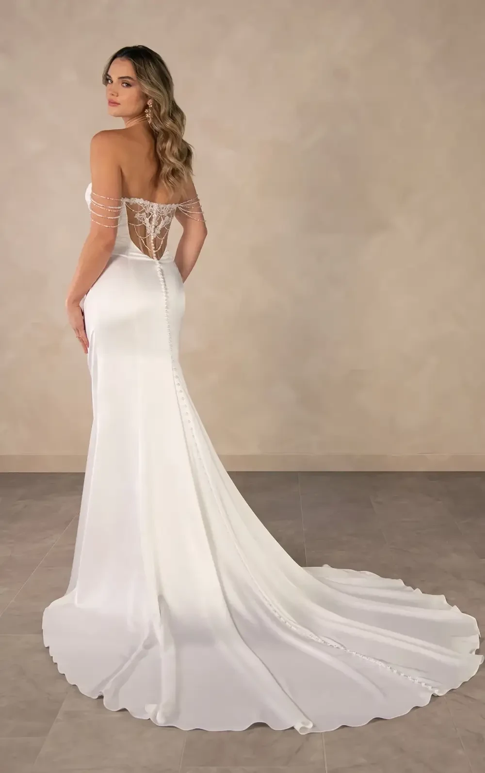 A woman in a elegant white gown with an intricate back design, facing away from the camera, with long hair styled in waves.