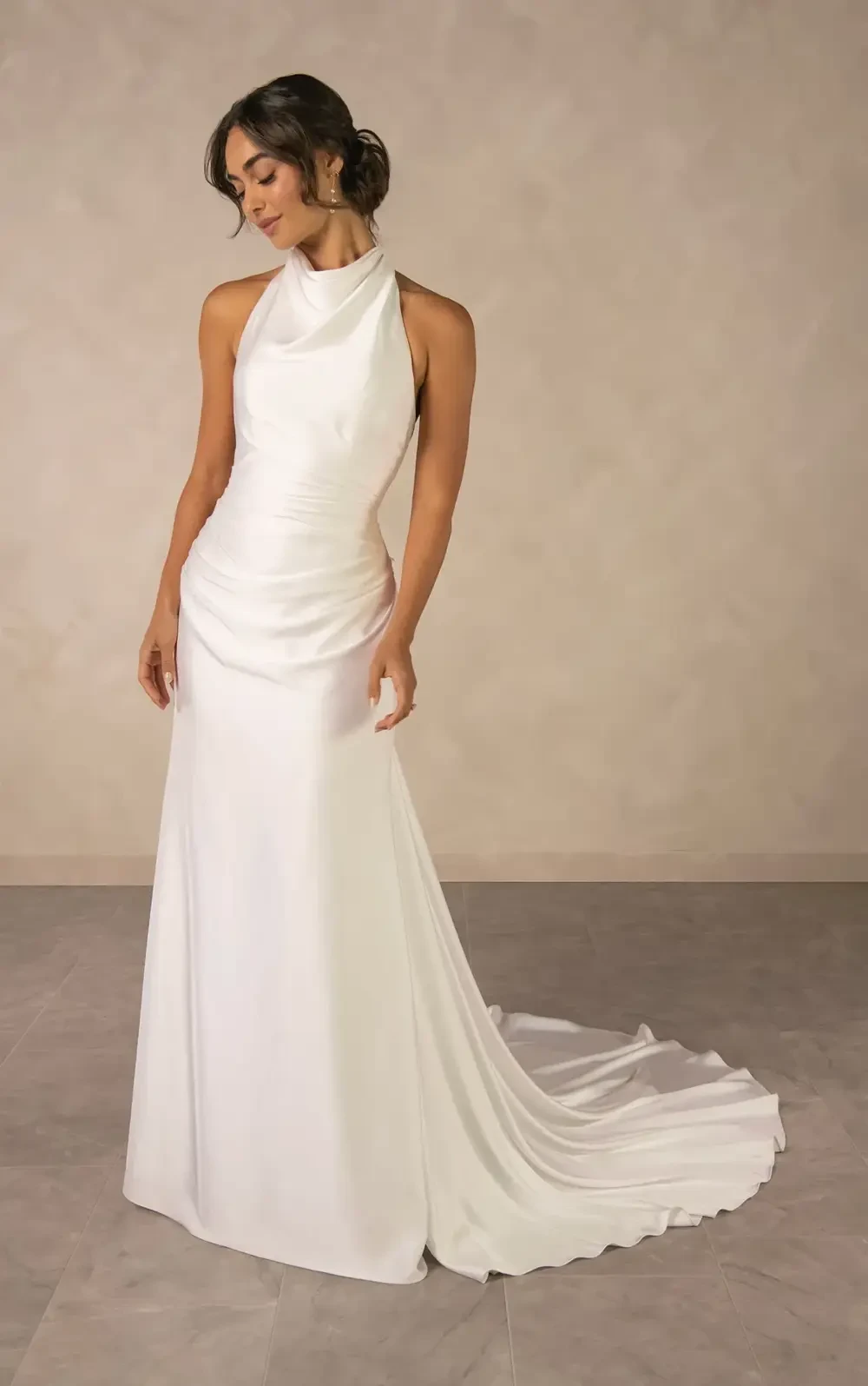 A model wearing a sleek, elegant white gown with a halter neck and a flowing train, standing gracefully against a soft, neutral background.