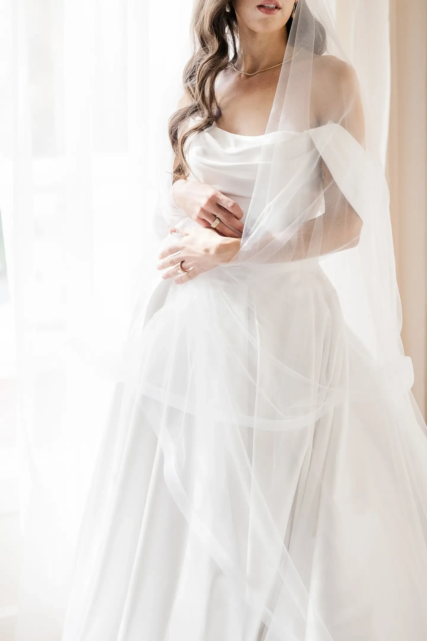 A bride wearing a flowing white dress and veil, standing by a window with soft natural light illuminating the scene.