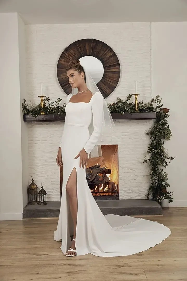 Model is wearing a sleek crepe gown with long sleeves, a square neckline, and a daring slit for the modern minimalist bride.