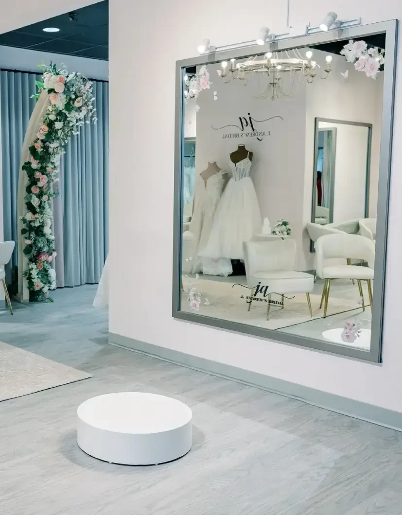 A stylish bridal boutique interior featuring a large mirror, a ceiling light fixture, and a floral decoration at the entrance. In the reflection, elegant wedding dresses on mannequins can be seen along with modern furniture.