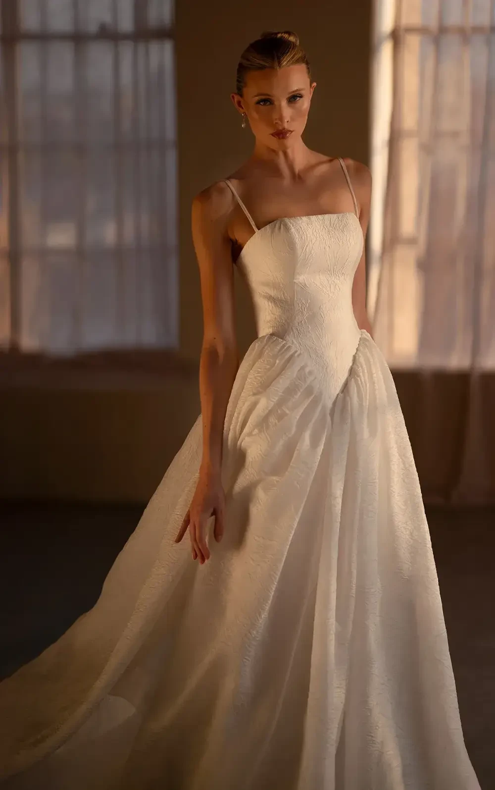 A model wearing a sophisticated white gown with a fitted bodice and a flowing skirt, posing in a softly lit room.