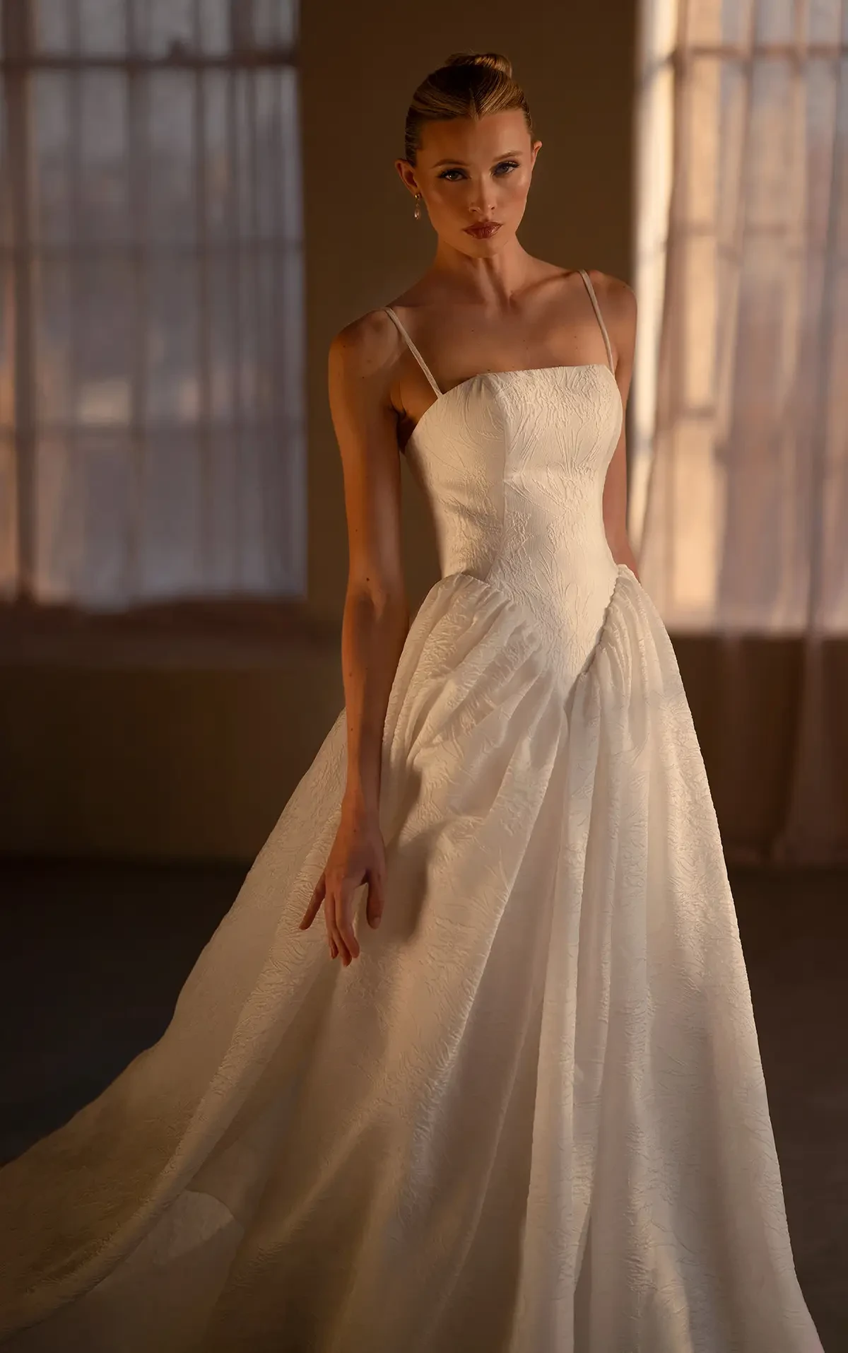 A woman in an elegant, textured white gown stands in soft lighting with a serene expression. The background features tall, draped windows, creating a calm, ethereal mood.