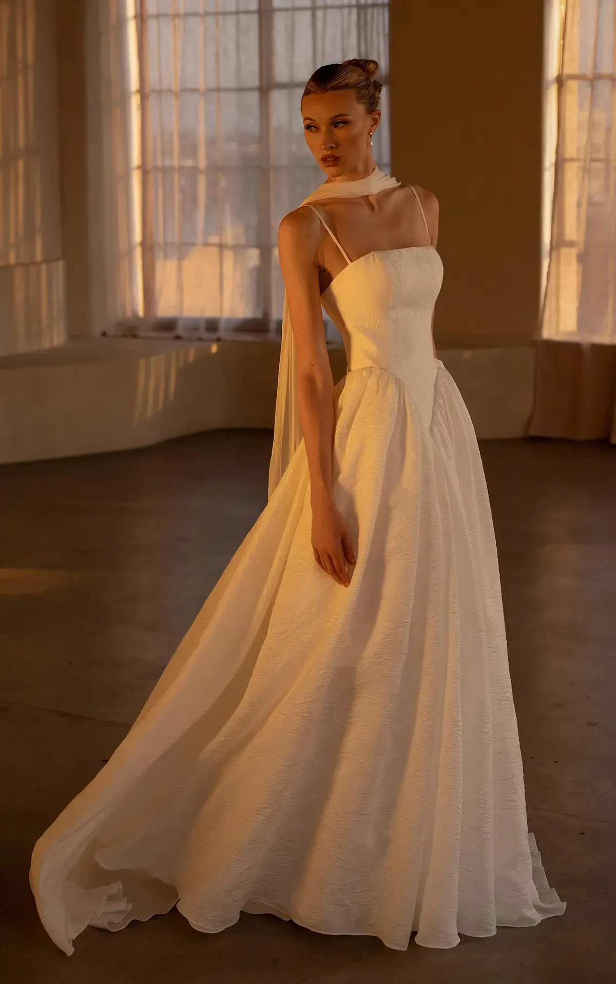 A woman in an elegant, flowing white gown stands gracefully in a dimly lit room. The soft lighting creates a warm, serene atmosphere.