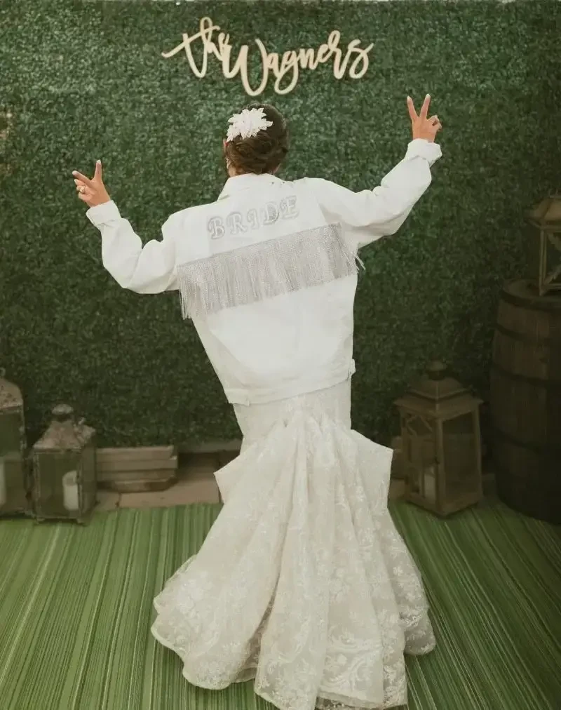 A person in a wedding dress with a long train and a white jacket, standing in front of a green backdrop that says 